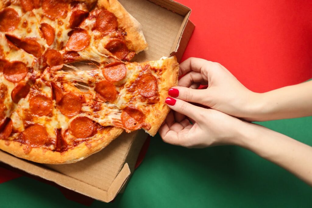 Close-up of a pepperoni pizza slice being pulled from the box on a vibrant red and green backdrop.